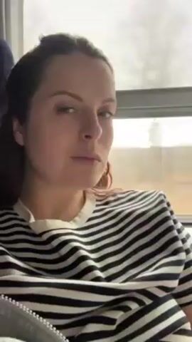 A light-skinned woman with brown hair is sitting, looking left out a window. She is wearing a striped black and white shirt. The background is blurred and shows an open field. The woman looks at the camera and back out the window.