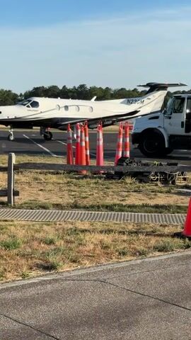 A white turboprop aircraft with the registration N236GM is parked on an asphalt surface, possibly an airport taxiway. The aircraft has black accents and a black tail. Several orange and pink traffic cones are positioned in front of the aircraft's left side. A white truck is partially visible to the right of the aircraft. In the foreground, a wooden fence and a strip of dry grass are present. The sky is blue with a few wispy clouds. The video appears to be filmed during daylight hours.