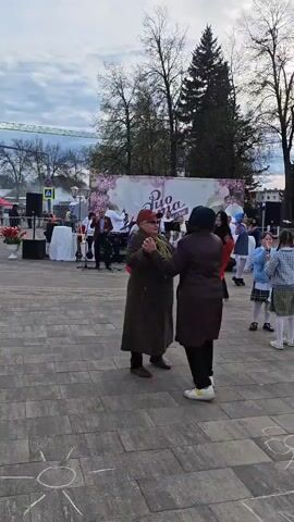A crowd of people are gathered at a brick-paved public square, with an ensemble performing on a stage in the background. Two older women in brown coats are slow dancing, while nearby a young girl in a pink jacket, blue jeans, and a white beanie walks towards the camera holding what looks like a bouquet of flowers.  Behind the young girl, a person walks past carrying a flag of Russia. A woman in all-black stands to the left, seemingly watching the activities.  A painted sun decoration is on the pavement in the foreground.  A sign above the stage reads "Duo Runa."  Trees surround the square and a construction crane is seen in the distance to the left of the stage. The sky is overcast.