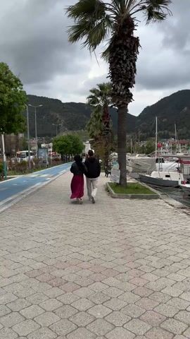 A couple walks away from the camera on a brick walkway lined with palm trees. The person on the left wears a black jacket and a long maroon skirt. She also appears to be wearing a black head covering. The person on the right wears a black hooded jacket and light-colored pants. A blue path, presumably for bicycles, runs parallel to the walkway on the left. Across the path is a street lined with buildings. On the right, boats are docked in a harbor next to the walkway. The sky is overcast, and mountains are visible in the background. A sign with the words "YASAL UYARI" is visible on the right.