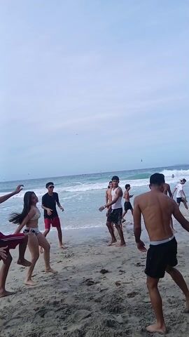 The video shows a group of young men playing volleyball on a beach. They are all wearing swim trunks, and some are shirtless. The beach is crowded with other people, and there are waves in the ocean. There's a hill with a group of people on it in the background. The weather appears to be pleasant.