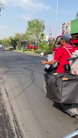 The video shows a street scene in Gelam, East Java. The time of day appears to be daytime, with bright sunlight casting shadows. The weather seems clear and sunny.

The primary subjects are a person riding a motorcycle and a large truck. The person on the motorcycle is wearing a red shirt, a grey cap, and is carrying a large, strapped package on the back of the scooter, along with two black saddlebags. The motorcycle is white.

The truck is red and laden with numerous green propane gas cylinders, which are strapped securely to the truck bed. The truck also has a cage-like structure at the back.

The scene depicts movement, suggesting that the vehicles are in transit. The motorcycle is in the foreground, moving away from the camera, while the truck is further back, also moving in the same direction. The road is paved, with a distinct curb and a grassy or dirt shoulder on the left side of the image. Trees are visible in the background, and a light pole stands tall on the right.

Visible text on the truck includes "PERTAMINA" and "GAS ALAM," indicating it is likely a Pertamina natural gas delivery truck. Another piece of text reads "UNTUK MASYARAKAT" which translates to "For the community."

The overall impression is a bustling street scene in a relatively urban or semi-urban area, likely during daylight hours, focused on transportation and delivery activities. There are no apparent indicators of specific emotions, but the presence of commercial vehicles and goods suggests a functional, everyday environment.