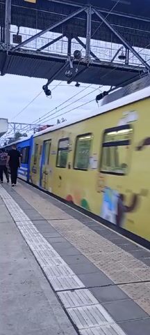 At an outdoor train platform on a cloudy day, a yellow train, partly covered with advertisements, travels from left to right. After it passes, a blue, white, and yellow train stops at the platform. Two men are seen standing on the platform. One walks toward the train. A gray metal structure covers the platform overhead.