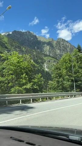 This is a first-person perspective video, likely recorded from a moving vehicle. The camera is pointed towards a majestic mountain landscape. In the foreground, a paved road with white lane markings is visible, bordered by a metal guardrail. Lush green trees, predominantly coniferous with some deciduous varieties, flank the road and cover the lower slopes of the mountains. A thin, white waterfall cascades down the rocky face of the mountain, surrounded by dense green forest. The sky above is a vibrant blue, dotted with a few scattered white clouds. The overall impression is one of a scenic drive through a mountainous, forested region on a clear day.