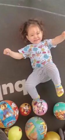 A young child, likely a toddler, is captured mid-air on a trampoline. The child is wearing a white t-shirt with a colorful cartoon print, grey striped pants, and yellow socks with a cartoon design. Their arms are outstretched to the sides, and their legs are bent, as if in the midst of a jump or bounce. The child's facial expression appears to be a mix of surprise and perhaps a bit of distress, with their mouth open and eyes wide.

Scattered on the trampoline surface are several colorful balls. These include a red and yellow ball, a blue and green ball with cartoon characters, and a larger, light blue inflatable ball with a cartoon owl design. A portion of the trampoline's black mat is visible, with a white logo or marking that appears to be part of the word "bounce."

The background suggests an outdoor setting, with glimpses of green grass and what looks like the metal frame of the trampoline. The lighting indicates it's daytime, though the exact time is difficult to ascertain. The overall scene depicts a playful, albeit slightly chaotic, moment of a child enjoying a trampoline with toys.