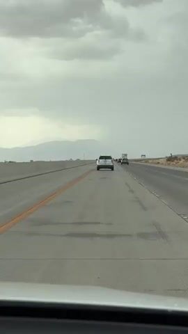 The video shows a view from inside a vehicle traveling on a highway. The sky is overcast with gray clouds. In the distance, there are mountains and desert-like terrain with sparse vegetation. Multiple vehicles are visible on the highway, including cars and a truck, all traveling in the same direction. The highway has multiple lanes and is divided by concrete barriers and painted lines. The road surface appears wet in some areas, suggesting recent rain. The perspective is from the driver's or front passenger's seat, looking forward.