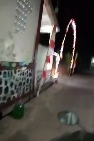The video, filmed at night, shows a person on a motorbike approaching a building with decorative lights. A silver car is parked to the right of the building and the motorbike stops beside it. A person is visible inside the car, and another person stands near the entrance of the building. The scene appears to be a residential area, with some vegetation visible in the background. The video is shaky and the lighting is dim, suggesting it was filmed with a handheld device in low light conditions.