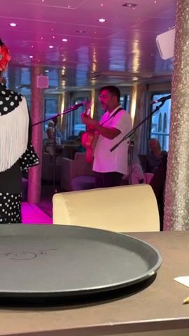 In this media, a man plays a guitar on a stage, illuminated by pink and white lights. He is wearing a white shirt and dark pants, with a microphone in front of him. To his left, a woman in a black and white polka dot dress with a white fringed shawl stands facing away from the camera. She is also wearing a red flower in her hair. In the background, several people are seated, watching the performance. The setting appears to be an indoor venue, possibly a restaurant or lounge, with a ceiling adorned with lights. The overall atmosphere is lively and celebratory. There is no visible text in the media. The scene suggests a musical performance is taking place.