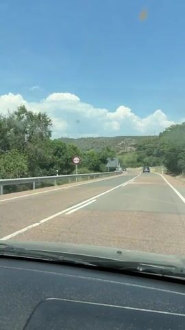 The view from the dashboard of a car driving on a winding two-lane road is shown in this video. The road is edged with guardrails and green trees. The sky is blue with some white clouds. A sign that says "no passing" is visible on the left side of the road. The car proceeds past another road sign. Other cars can be seen on the road ahead. A large white sign appears to be a road marker showing directions for Jérica. A mountain covered with sparse trees is in the background.