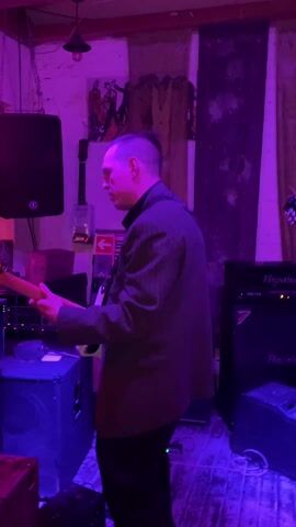 A man in a suit plays a bass guitar in a room bathed in blue and purple light. The walls are unfinished and a black speaker is mounted on the wall to his left. Behind him and to his right are guitar amps. Another man, with longer hair, plays DJ equipment next to him. The room looks like a music venue, and the two are presumably performing.