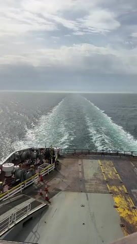 The video shows the view from the rear deck of a large ship traveling on the ocean. The ship is leaving a long, turbulent wake of frothy white water that extends to the horizon. The deck is metal, with worn yellow paint indicating lanes. There are some mechanical structures and equipment toward the left side of the deck. Orange traffic cones are visible, suggesting ongoing operations. The sky is overcast with a mix of gray and white clouds, and the ocean is a grayish-blue. The horizon is somewhat indistinct due to the atmospheric conditions.