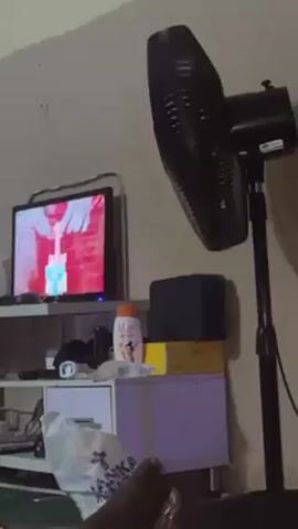 A dimly lit room featuring a television displaying a close-up image of hands manipulating a green and white object with red accents. A black standing fan is positioned next to a white and lavender-colored cabinet holding items like lotion, boxes, and a small black speaker. A white plastic bag sits on the lower shelf of the cabinet, and a foot is visible in the bottom left of the frame.