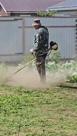 A person wearing camouflage clothing and a hat is trimming overgrown grass with a string trimmer in a yard. Dust is flying up from the ground as the trimmer operates. The person is standing near a metal fence and a building.