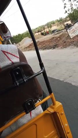 The video, shot from the passenger seat of a yellow auto-rickshaw, shows a ride through a rural or semi-rural area. The driver, visible in the rickshaw's mirror, wears sunglasses and a white jersey with a red and black graphic. There are at least two passengers: one in a dotted white shirt, and another in a blue shirt. The camera is focused on the back of the front seat, which is dark brown and has metal fasteners. The rickshaw travels along a paved road, passing a construction site with piles of sand, bricks, and building materials. The background shows greenery and scattered buildings.  The footage appears to be taken during the daytime.