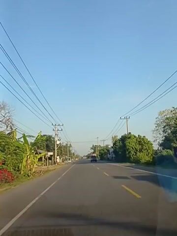 The video shows a view from inside a vehicle traveling on a paved road. The road has yellow lane markings. On both sides of the road, there are power lines supported by poles, some of which have transformers. There are also trees, vegetation, and a few small buildings or structures lining the road. In the distance, there are a few vehicles, including a truck and some motorcycles. The sky is clear and blue, suggesting it is daytime. The overall scene depicts a rural or suburban setting with ongoing road traffic and infrastructure.