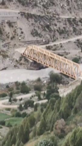 This aerial video showcases a remote, mountainous landscape in Tandi, India. The primary object of focus is a long, yellow truss bridge spanning a wide, murky river. The bridge appears to be made of steel and is supported by a complex network of diagonal beams. The river below is fast-flowing, with visible currents.

The surrounding terrain is rugged and steep, characterized by rocky slopes and sparse vegetation. The slopes are dotted with scattered trees and shrubs, with some patches of green, terraced fields visible in the lower portions of the frame. A winding dirt road can be seen running alongside the river and up the mountainside. Another road is visible higher up on the slope.

The scene suggests a remote and somewhat undeveloped area, likely far from urban centers. The natural environment is dominant, with the bridge serving as a vital piece of infrastructure connecting the two sides of the valley. The time of day appears to be daytime, with natural lighting illuminating the scene. The weather seems overcast or hazy, as the light is diffused. No people are visible in the footage. There is no visible text in the video. The overall mood is one of natural grandeur and the engineering feat of the bridge in such a challenging environment.