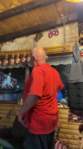 The video shows an older man in a red polo shirt standing indoors in a wooden cabin. He turns to face the camera, smiles, and then turns away again. The camera momentarily moves upward to show more of the room. In the background, there is a fake fireplace on a TV screen, a mounted bird of prey, and a young boy sitting in a wicker chair and looking at his phone. The cabin is decorated with folk art and rustic items. There are copper pots hanging above the window. The ceiling is wooden.