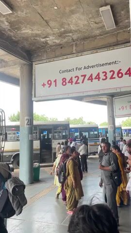 The video depicts a bustling scene at a bus station. Several people are waiting near a bus. An advertisement banner for advertising opportunities is visible. The bus is labeled "Himachal Path Parivahan Nigam Mandi". People are boarding the bus, and some are standing around with luggage. The atmosphere is typical of a busy transportation hub.