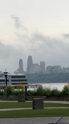 The video pans across a waterfront park with a cityscape in the distance. In the foreground, there is a grassy lawn with two dark stone sculptures. A low wooden planter box filled with green plants and yellow flowers sits behind the lawn. Beyond the planter box is a body of water. On the other side of the water, there is a treeline and a dense urban skyline with several tall buildings, some with distinctive architectural features. The sky is overcast with clouds. The camera moves to the right, revealing a lamppost and a bench in the park. The cityscape remains visible throughout the pan.