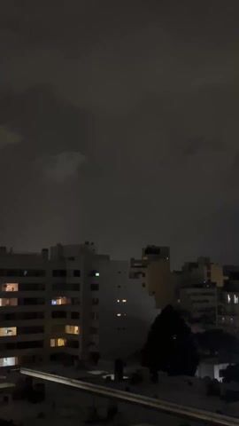 The scene is a nighttime cityscape.  Several buildings are visible, silhouetted against a dark, cloudy sky.  Some windows in the buildings are lit.  A tree can be seen in the foreground. There is the sound of distant traffic.