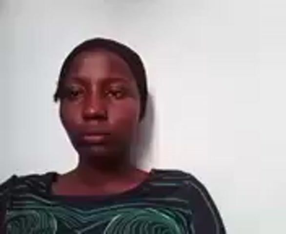 A dark-skinned woman with a head covering looks directly at the camera. She touches her face as she speaks. She is wearing a dark, patterned shirt. The background is a plain white wall.