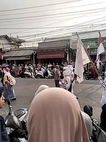 A daytime outdoor scene shows a street parade. In the foreground, partially obscured, are the backs of several people wearing headscarves. They are on a paved street, with motorcycles parked on the left. In the middle ground, a group of individuals in white uniforms with white head coverings are marching. Some are holding flags: one white flag with text and an Indonesian flag. Behind them, more people in white uniforms are marching, and to the right, a group of individuals in white uniforms and hats are also marching. Spectators are gathered on the sidewalks on both sides of the street, many on motorcycles. Buildings line the street, some with signs that are partially visible. The sky is overcast.
