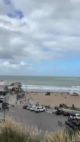 The video presents an elevated view of a beach with a sandy shoreline, a body of water extending to the horizon, and a sky partially covered with clouds. Several people are scattered along the beach, and there are also umbrellas set up. A road is visible in the foreground, lined with parked cars. Tall grass grows nearby, partially obstructing the view. Beyond the beach, a harbor structure can be seen extending out into the sea.