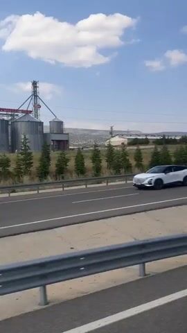 The video shows a side view of a large industrial facility with multiple metal silos and a tall tower structure. The facility is situated on a slight incline, with a line of evergreen trees separating it from a two-lane road in the foreground. The road appears to be an asphalt highway, and a shoulder with dry grass and some rocks is visible on the right side. The sky is blue with scattered white clouds, and the landscape in the background is hilly and sparsely vegetated. The perspective suggests the video is being filmed from a moving vehicle. There are no people visible in the video.