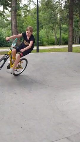 The video captures a boy riding a yellow bicycle in a skate park. He is wearing a black t-shirt and black shorts. A water bottle is attached to the handlebars of his bike. The skate park is made of concrete and is surrounded by trees and lush greenery, suggesting an outdoor setting during the day. The weather appears to be pleasant and overcast, as indicated by the soft lighting.

In the background, another person is also seen with a bicycle, partially obscured by trees, and a blurry figure on a bicycle is visible in the distance, suggesting other people are present and engaging in similar activities. The boy is actively riding his bike, navigating the curves of the skate park, and appears to be enjoying himself. His posture and the slight turn of his head suggest he is focused on his ride and possibly interacting with the environment around him. The presence of a skate park implies a recreational area designed for activities like skateboarding, biking, and rollerblading. The scene evokes a sense of youth and active leisure in a park-like environment.