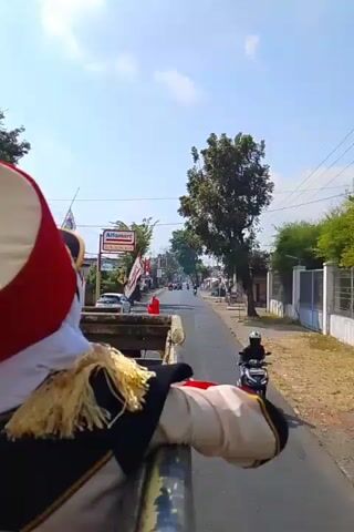 The video shows a first-person perspective from a moving vehicle, possibly a truck bed, as it travels down a paved road. The camera is focused on a person wearing a marching band uniform, including a red and white hat, a black jacket with gold trim, and a white scarf. The person is leaning over the side of the vehicle, facing forward. The road is lined with buildings, trees, and utility poles. Indonesian flags are visible on some of the buildings. There are a few other vehicles, including motorcycles, on the road. The sky is blue with some scattered clouds. The overall scene suggests a parade or a procession.