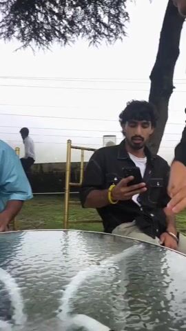 A man with dark hair and a beard, wearing a white t-shirt and a black open button-up shirt, sits at a glass-top table in an outdoor setting. He holds a smartphone in his left hand and appears to be interacting with it. Another person, partially visible and wearing a black long-sleeved shirt and light-colored pants, stands to the right of the frame, holding what appears to be a pair of scissors. The background shows a lush green lawn, trees, and a hedge. The sky is overcast. The man at the table looks towards the person with the scissors.