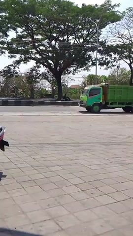 A bright green truck is driving on a paved road. The truck has a large cargo bed, which is also green and appears to be empty. There are large trees with full green foliage in the background, and the sky is clear and blue. The road is paved with large, square gray pavers. The video is shot from a low angle, looking up at the truck and the surrounding environment. The truck is the main subject of the video.