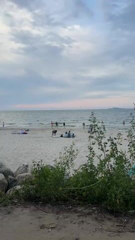 This media file depicts a serene beach scene in Kulikovo, Russia. The time of day appears to be late afternoon or early evening, as suggested by the soft, diffused light and the pale, cloudy sky. The weather is calm, with gentle waves lapping at the shore.

The foreground is dominated by a sandy beach with some vegetation and rocks. In the mid-ground, several people are enjoying the beach. Some are seated on the sand, possibly picnicking or relaxing. Others are wading or swimming in the shallow waters of the sea, indicating a leisurely activity. A few individuals are standing near the water's edge. In the distance, a large ship is visible on the horizon, adding a sense of scale to the vastness of the sea.

The overall mood of the scene is tranquil and peaceful, with a sense of a pleasant, relaxed day at the beach. The people appear to be enjoying their time, and the natural environment contributes to a feeling of calm. There is no visible text in the media file.
