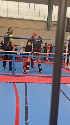 Here's a description of the video:

The video shows a kickboxing match in a boxing ring. Two fighters, wearing protective gear, stand in the ring. A referee in a lime green and black tracksuit is addressing one of the fighters, adjusting the fighter's helmet. A crowd of spectators watches from outside the ring. The ring has blue and red mats. The room has a high ceiling and is illuminated by overhead lighting. The camera's view is partially obstructed by a vertical pole.