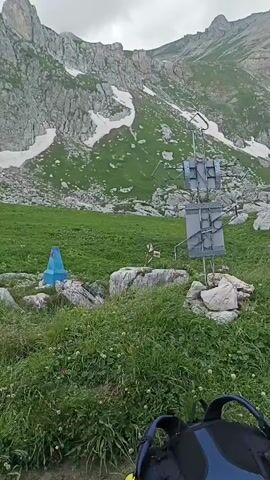 A video of a mountainous landscape shows a grassy field with a metal structure in the center. The structure has two panels that appear to be signs or displays. To the left of the structure is a blue and orange marker. In the background, a large mountain range is visible, with patches of snow on its slopes. The sky is overcast. The bottom of the video shows the top of a black and blue backpack.
