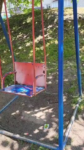 This media file features an outdoor scene of an empty swing set. The swing set has a metal frame painted blue and red chains supporting a red metal seat with blue paint splattered on it. The ground beneath the swing set is dirt and dry grass, with some scattered leaves. In the background, a grassy slope is visible, with a building with white windows and a tiled roof at the top. Sunlight dapples the scene, suggesting it is daytime, likely with clear weather. There is no visible text in the image. The overall mood of the image is somewhat melancholic due to the emptiness of the swing and the worn appearance of the playground equipment. The location context is Zolotonosha, Ukraine.