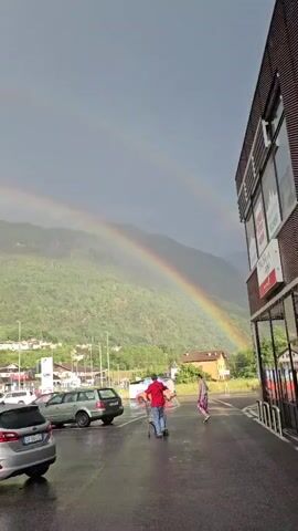 The video begins with a wide shot of a cloudy sky with a double rainbow visible against a backdrop of a lush green mountain. In the foreground, a parking lot is wet, suggesting recent rain. Several cars are parked in the lot, and two people are walking away from the camera, one using a walker. To the right, a modern building with large windows is visible, and further in the distance, houses and smaller buildings are scattered across the mountainside. The scene is tranquil, with the natural beauty of the rainbow and mountains contrasting with the man-made structures and the signs of everyday life.