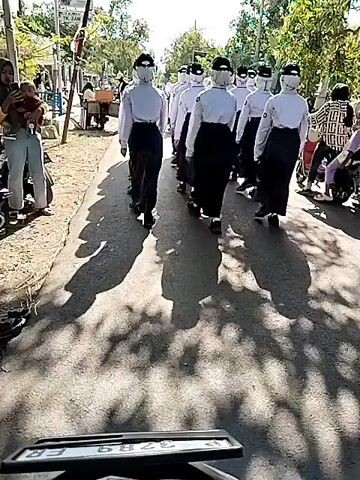 A group of people dressed in white shirts, black pants, and face shields are walking down a paved road in a single file line. They are walking away from the camera. The road is lined with trees and some buildings. There are other people on the side of the road watching them. The sun is shining and there are shadows on the road. The video appears to be shot from a moving vehicle.