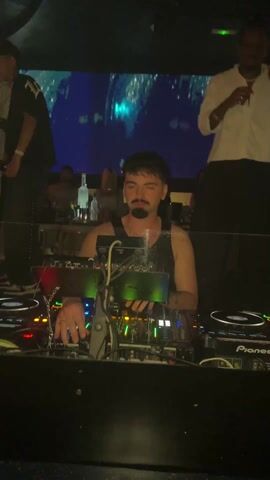 The video shows a DJ with a dark mustache and beard, wearing a black tank top, operating DJ equipment in a dimly lit nightclub. Behind him, several people are standing, including a man in a white shirt and black pants who is actively dancing with his arms outstretched. The background features a screen displaying abstract blue and white visuals. There are also red inflatable tubes positioned near the DJ setup. The scene conveys a lively atmosphere with people enjoying themselves in a club setting.
