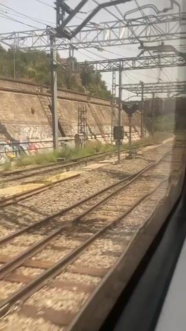 The video shows a view from inside a moving train, looking out a window. The train tracks are visible in the foreground, with ballast and gravel. To the left, there is a retaining wall with graffiti on it, and a grassy slope with vegetation. Overhead, there are electrical wires and supports for the catenary system. Further down the tracks, a railway signal can be seen. The train is moving on a curved track, and the scenery is blurred due to the motion. The lighting suggests it is daytime. There are no people or animals clearly visible within the frame.