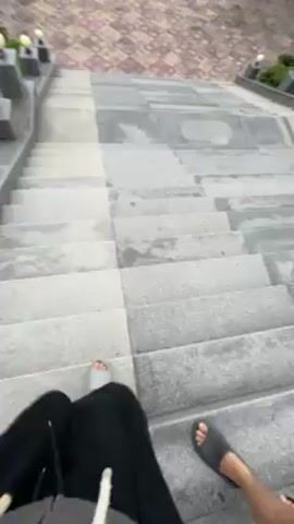 The video shows a first-person perspective of two people walking down a large stone staircase. The person filming is wearing black pants and has a bare foot visible in a sandal. Another person beside them is wearing sandals. At the bottom of the stairs, there is a large paved area with a checkered pattern and ornamental lights on either side of the stairs. Beyond the paved area, there are trees, power lines, mountains in the distance, and an overcast sky.
