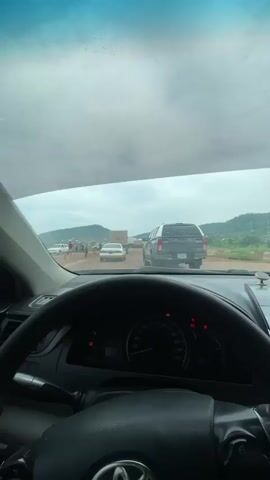 The video captures a traffic scene, likely on a road under construction or with poor road conditions. The first frame shows the interior of a car looking through the windshield, with a dashboard and steering wheel visible. Outside, several cars are stopped in traffic. The road appears to be dirt or gravel, and there are some distant hills or mountains. The second frame provides a side view from inside a vehicle, looking out the side window. A large red shipping container truck is parked on the side of the road, with a blue car and a red pickup truck nearby. The ground is muddy and there are puddles of water. The third and fourth frames continue to show the side view, with the red pickup truck and a blue van visible, along with more muddy patches and puddles on the road. The sky is overcast with clouds. The overall impression is of a slow-moving or halted traffic situation due to difficult road conditions.