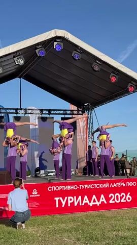Here's a description of the video:

Acrobatic performers are giving a display on an outdoor stage. There are three pairs of people, with the bottom person of each pair holding the other person upside down. The base performers are wearing purple tops and pants, while the top people are in purple and yellow outfits. There are other people in purple uniforms standing to the side of the stage. A woman is crouched at the front, taking pictures of the performance with her smartphone. The stage is covered by a white canopy and has several stage lights. Behind the stage is a red banner with the text "СПОРТИВНО-ТУРИСТСКИЙ ЛАГЕРЬ ПФО ТУРИАДА 2026". Some people in military uniforms are seen standing behind the stage. The sky is blue and clear. The setting seems to be outdoors, with greenery visible in the background. One of the acrobats dismounts and catches the other, then all of them do the same.