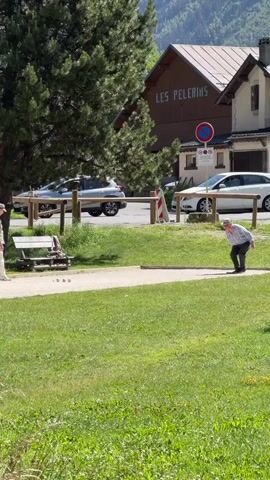 The video shows an outdoor scene in what appears to be a village or town. In the foreground, there is a grassy area. A gravel path runs through the middle of the frame, and on this path, there are three metal balls. To the left of the path, there is a park bench with some equipment on it.

On the right side of the path, a man is playing a game that involves throwing balls. He is wearing a hat, a plaid shirt, and dark pants. He is crouched down, in the motion of throwing a ball.

In the background, there is a tall evergreen tree to the left, and buildings to the right. One building has the sign "LES PELERINS" on it. There is a street behind the path with cars parked and driving on it. In the far background, there are mountains. The weather appears to be sunny.