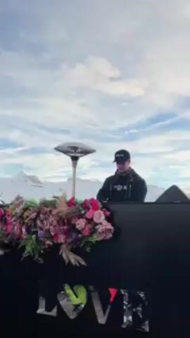 The video captures an outdoor party scene atop a snowy mountain, likely a ski resort. In the foreground, a DJ in a black jacket and baseball cap stands behind a booth decorated with pink and purple flowers and the word "LOVE" in mirrored lettering. A large sound system broadcasts music. The background reveals a breathtaking vista of snow-covered peaks under a partly cloudy blue sky. Several people, dressed in ski attire including jackets, headbands and helmets, are milling around, many holding drinks. The scene suggests a lively après-ski celebration with music and a stunning mountain backdrop.