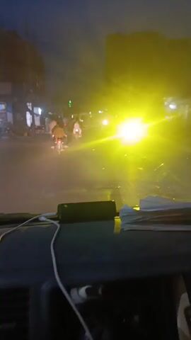 The video shows a view from inside a vehicle driving down a street at dusk or night. Several people are visible riding bicycles and motorcycles, and a few are walking on the sidewalks. Buildings with storefronts line the street, some with their gates closed. A silver car passes on the left. The street is lit by the vehicle's headlights and streetlights. The vehicle's dashboard is visible, along with a charging cable and some papers. The overall atmosphere suggests a typical evening scene in a busy urban area.