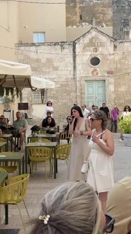 A female singer performs live in an outdoor setting in Monopoli, Italy. She stands centrally, holding a microphone and wearing a light-colored, buttoned dress and heels. Numerous people are present, forming an audience. Some are seated at green-yellow cafe tables with white umbrellas in the foreground and to the left, while others are standing and watching in the background.

The performance takes place in a paved square or street, flanked by old stone buildings. Directly behind the singer is a historic stone building, possibly a chapel or church, featuring a light blue wooden double door, a small circular window above the door, and a cross on its roofline. A small, unreadable red sign is affixed to this building. The overall atmosphere appears relaxed and engaging, with onlookers enjoying the music on a bright, clear day.