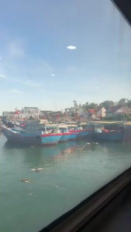 A clear sky with some scattered clouds is visible above a harbor filled with various fishing boats. The boats are moored near a coastline lined with buildings. The water in the harbor appears calm with some debris floating on the surface. The perspective is from inside a vehicle, likely a train or bus, looking out through a window. The window frame is visible in the foreground, suggesting movement or a continuous view.