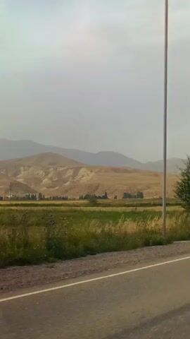 The video is taken from a moving vehicle, likely a car, looking out the side window. It shows a rural landscape with a paved road in the foreground. On the left side of the road, there is a field with green vegetation, possibly crops or grass, bordered by a dirt or gravel shoulder. Beyond the field, there are rolling hills with dry, light brown vegetation. The sky is overcast and hazy. A few scattered trees and bushes are visible along the side of the road and in the distance. There are also a few utility poles and streetlights along the road. In the distance, a few cars can be seen driving on the road. The overall impression is of a somewhat barren but open landscape.