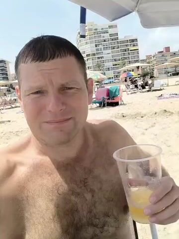 A man is taking a selfie on a beach. He is shirtless and holding a plastic cup with a yellow drink in it. He is smiling at the camera. In the background, there are beach chairs, umbrellas, and buildings. The sky is blue and there are a few clouds. The man appears to be on vacation.